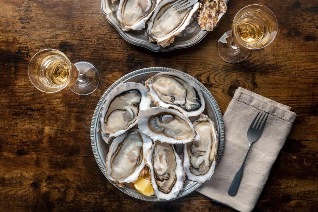 Plate of fresh raw oysters on the half shell served with lemon wedges and glasses of white wine on wooden table