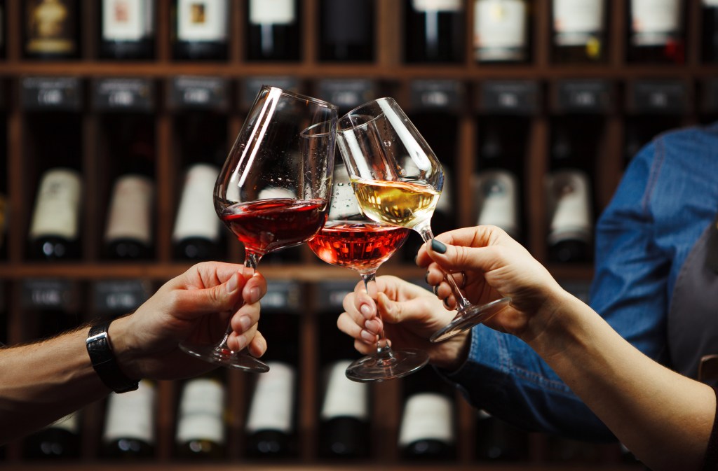 Three people clinking glasses of red, rosé, and white wine in a wine shop with shelves of bottles in the background