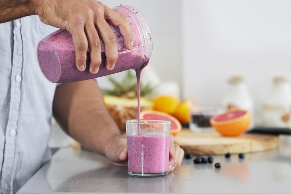 Hand pouring a purple berry smoothie into a glass with pineapple, grapefruit, and blueberries on a kitchen counter