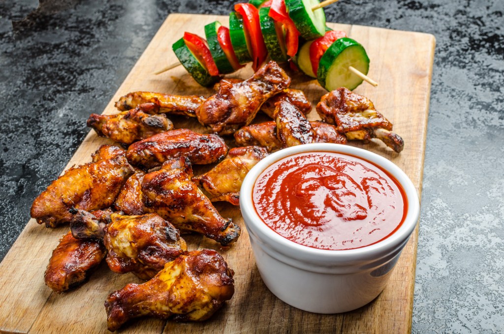 Glazed BBQ chicken wings served with a bowl of barbecue sauce and grilled vegetable skewers on a wooden board