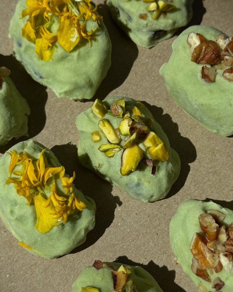 Green coated Medjool dates topped with pistachios pecans and yellow edible flowers on parchment paper