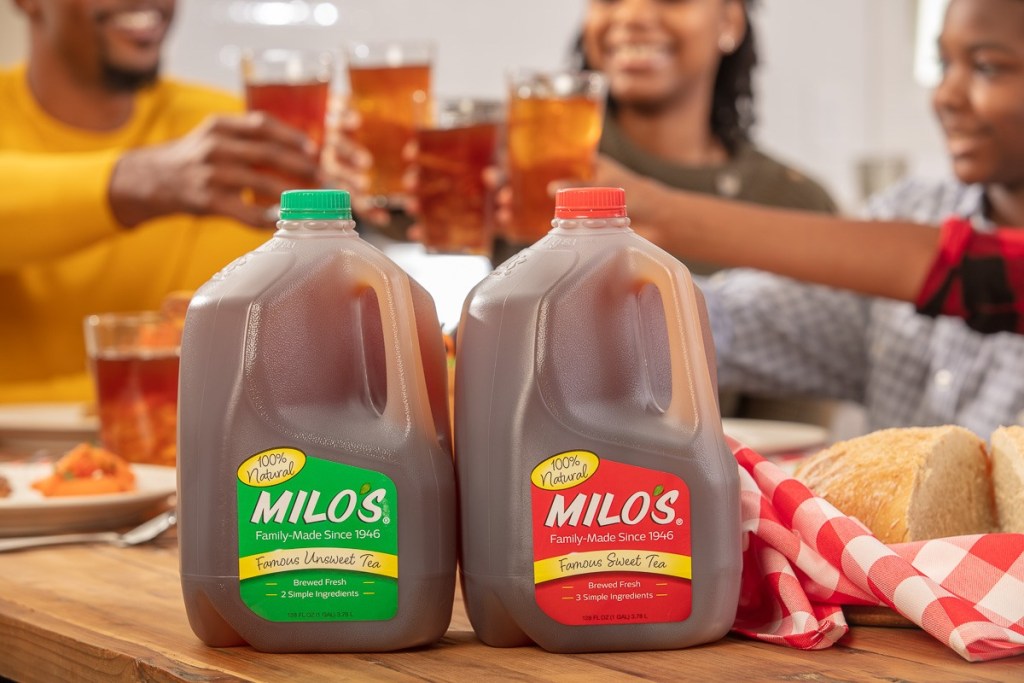 Gallon jugs of Milos sweet tea and unsweet tea on table with family enjoying drinks in background