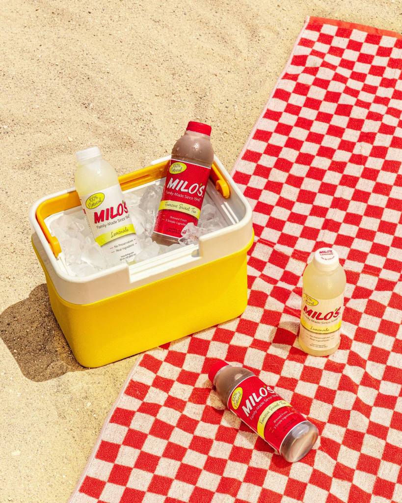 Milos sweet tea and lemonade bottles chilling in ice filled cooler on sand with red checkered picnic blanket