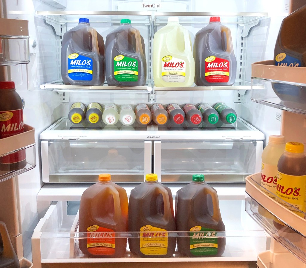Multiple gallon jugs and bottles of Milos tea and lemonade arranged inside a refrigerator on shelves
