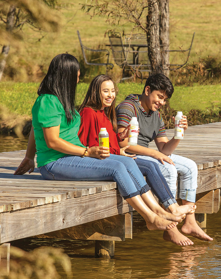 Three people sitting on a wooden dock holding bottles of Milos tea and lemonade enjoying a sunny day by the water