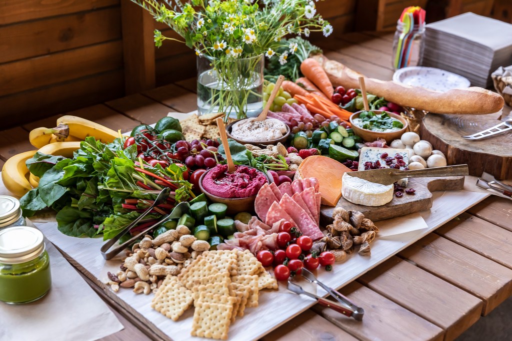 Colorful grazing board with fresh vegetables fruits cheeses crackers dips and cured meats arranged on a wooden table outdoors