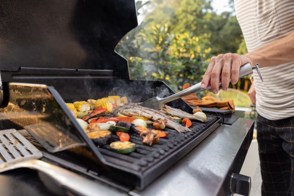 Person grilling whole fish with corn, zucchini, peppers, and onions on an outdoor barbecue grill