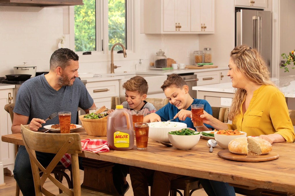 Family enjoying a meal together with glasses and gallon jug of Milos sweet tea on a wooden dining table