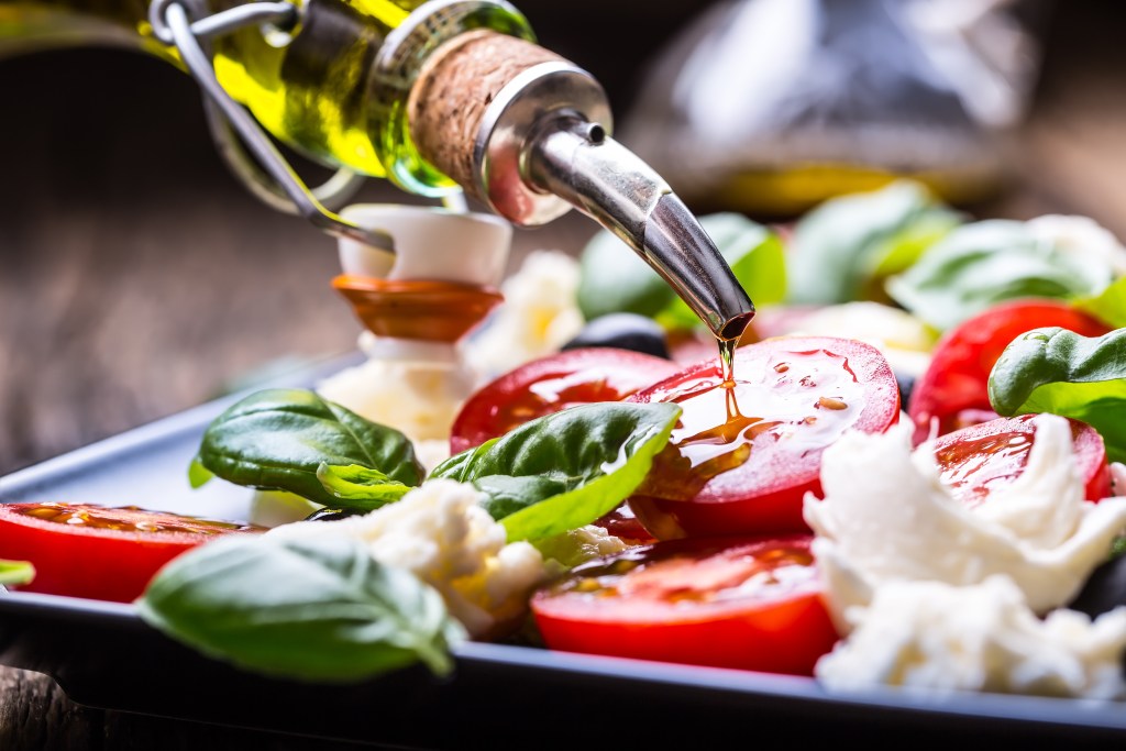 Olive oil being poured over a Southern caprese salad with sliced organic grape tomatoes fresh basil and mozzarella cheese