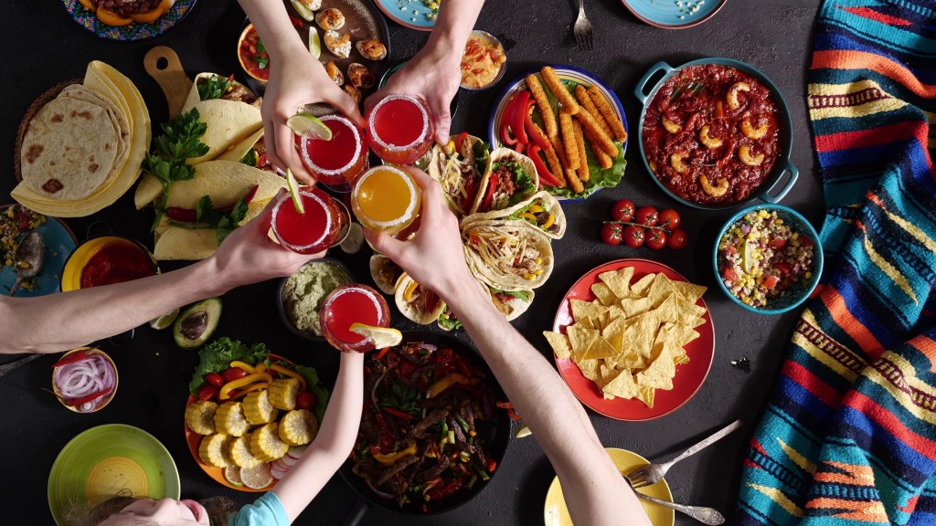 Overhead view of friends toasting margaritas over a colorful Cinco de Mayo spread with tacos nachos chips salsa guacamole and fajitas