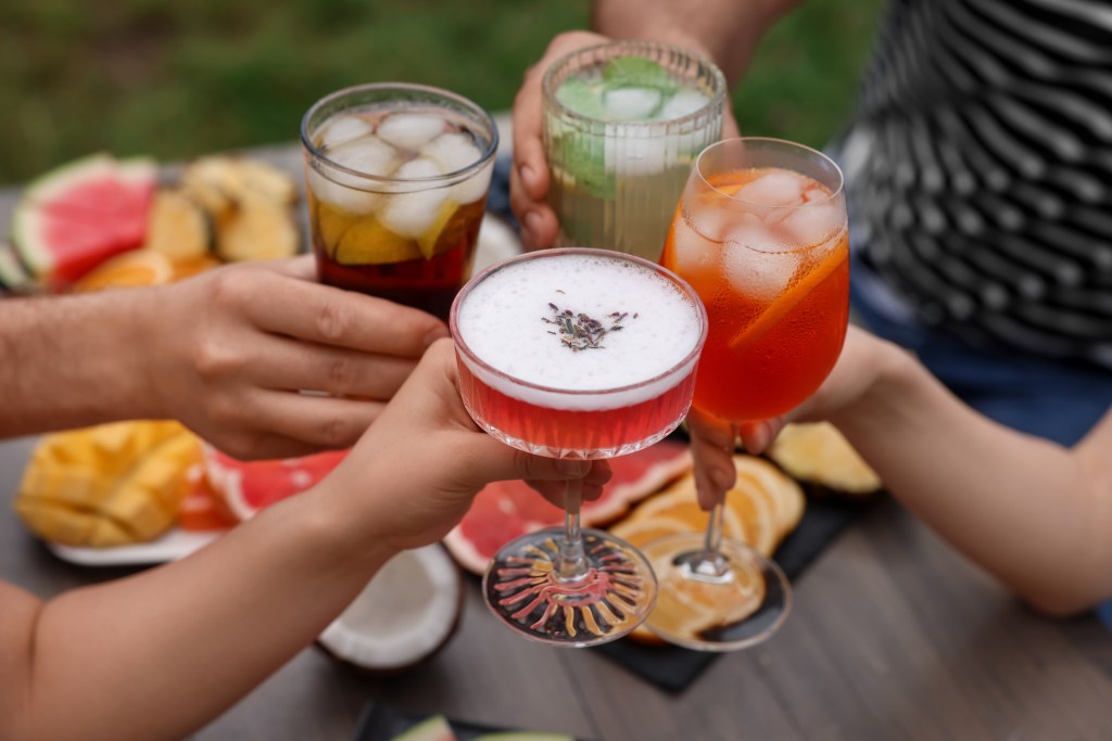 Friends toasting colorful summer beverages with fresh fruit and iced drinks at an outdoor table with watermelon, citrus, and tropical fruit