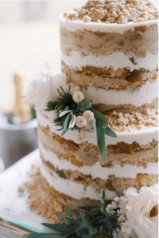 Two-tier naked wedding cake with white frosting layers, crumble topping, and floral greenery accents on display at a celebration