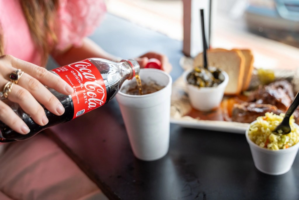 Person pouring Coca-Cola Original Taste from a 2-liter bottle into a cup while enjoying a meal, promoting a free Coca-Cola product offer at Piggly Wiggly Birmingham