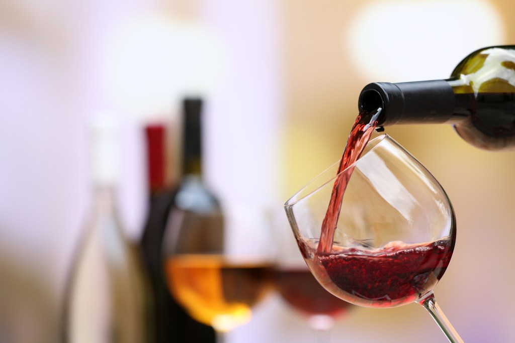 A bottle pours red wine into a stemmed wine glass with blurred bottles of red and white wine in the background, illustrating wine tasting and varietal selection.