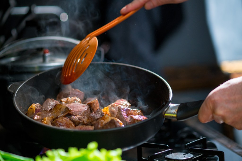 Fresh beef cubes searing in a skillet on a stovetop while being stirred with a cooking utensil, demonstrating safe meat cooking and proper food preparation.
