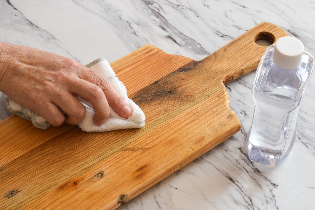 Hand cleaning a wooden cutting board with a cloth and cleaning solution to prevent cross-contamination and maintain safe food preparation practices.