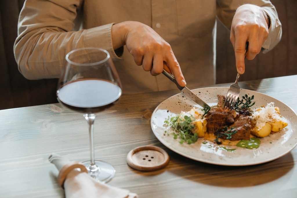Person cutting a gourmet meat dish served with roasted potatoes and vegetables alongside a glass of red wine in a fine dining restaurant