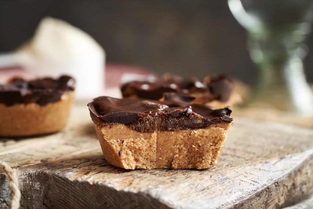 Close-up of chocolate peanut butter dessert cups featuring a buttery cookie crust, creamy peanut butter filling, and a rich chocolate topping on a rustic wooden board.