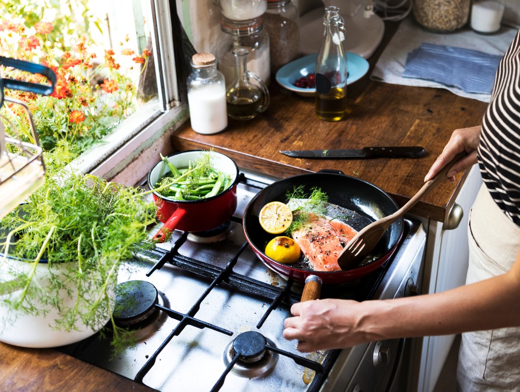 Person cooking a pan-seared salmon fillet with lemon and fresh herbs on a stovetop beside a pot of green vegetables in a home kitchen