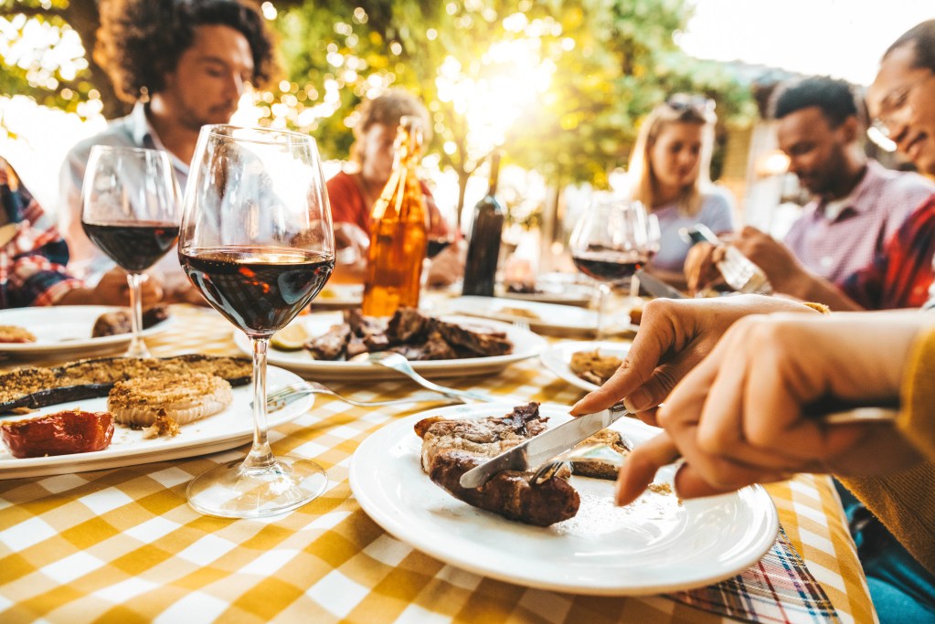 Friends enjoying an outdoor meal with grilled steak, glasses of red wine, and shared dishes at a sunlit dining table