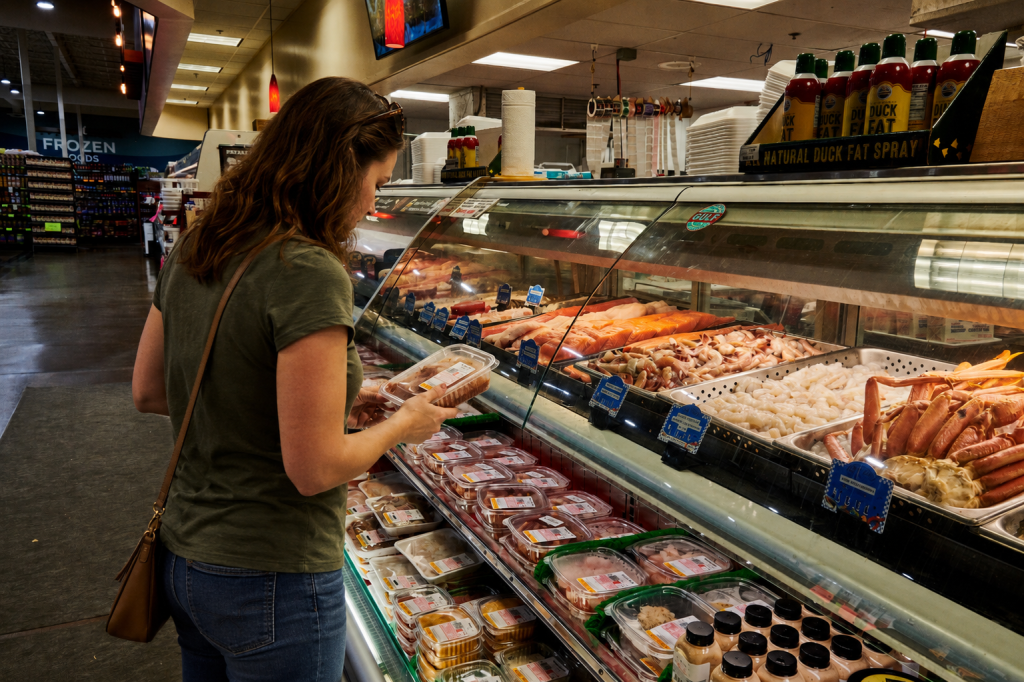 Customer examining packaged meat labels while shopping at the fresh meat counter inside Piggly Wiggly Birmingham, comparing cuts and product information before making a purchase.