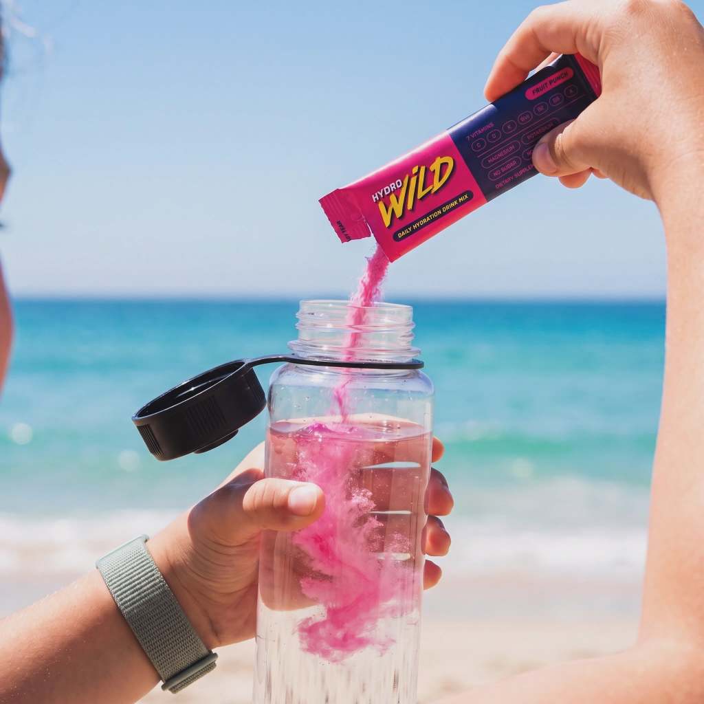 HydroWild Fruit Punch hydration drink mix being poured from a single-serve stick into a water bottle at the beach