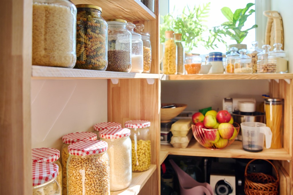 Organized family pantry with glass jars of rice, pasta, grains, lentils, and other staples alongside fresh apples and eggs.