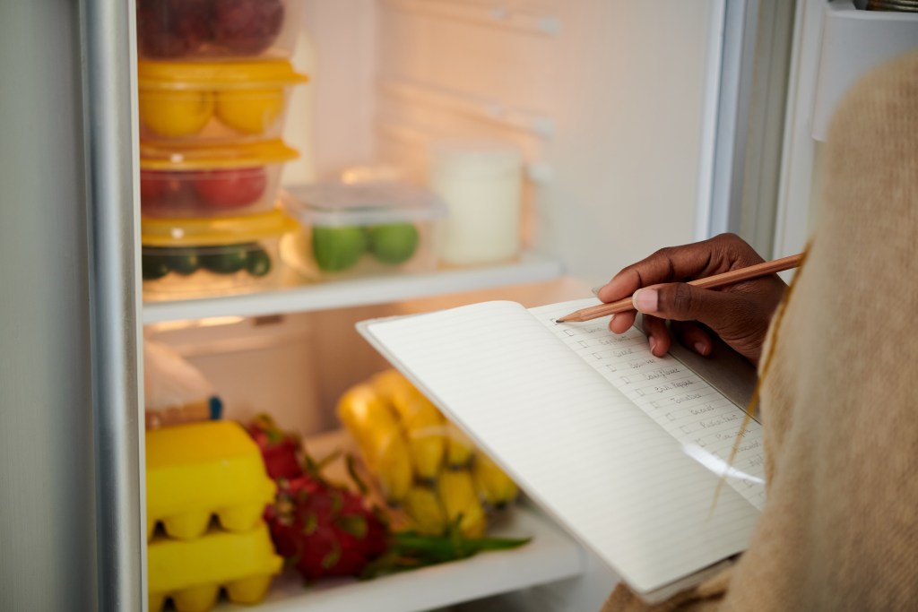 Person checking a grocery list while reviewing food, produce, and staples already stored in the refrigerator