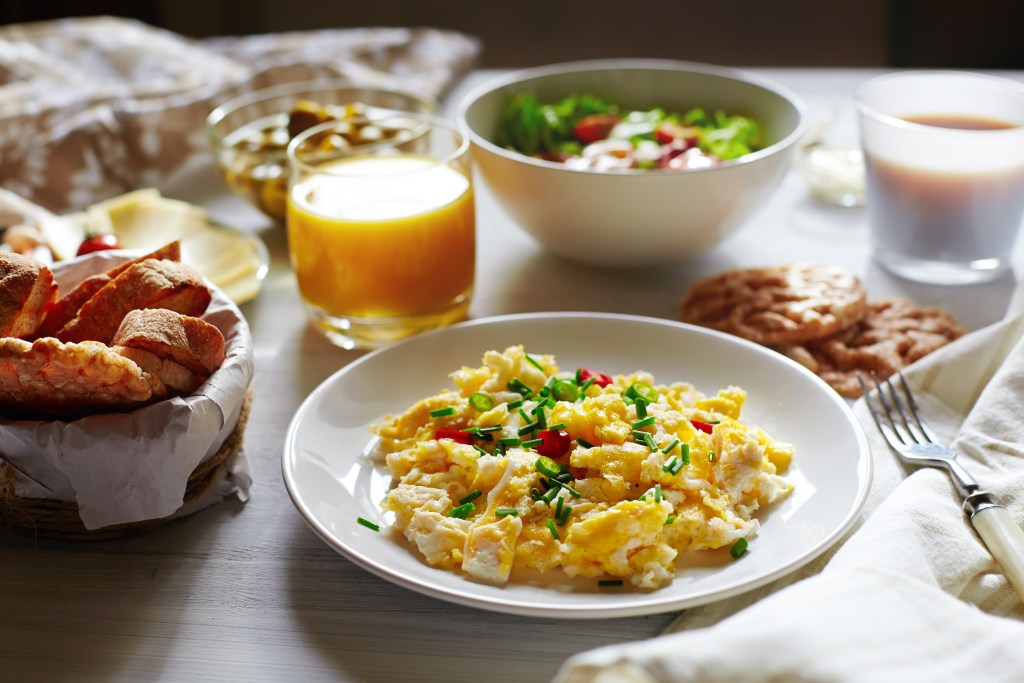 Scrambled eggs with chives served alongside bread, fruit, juice, and other breakfast foods for a busy school morning