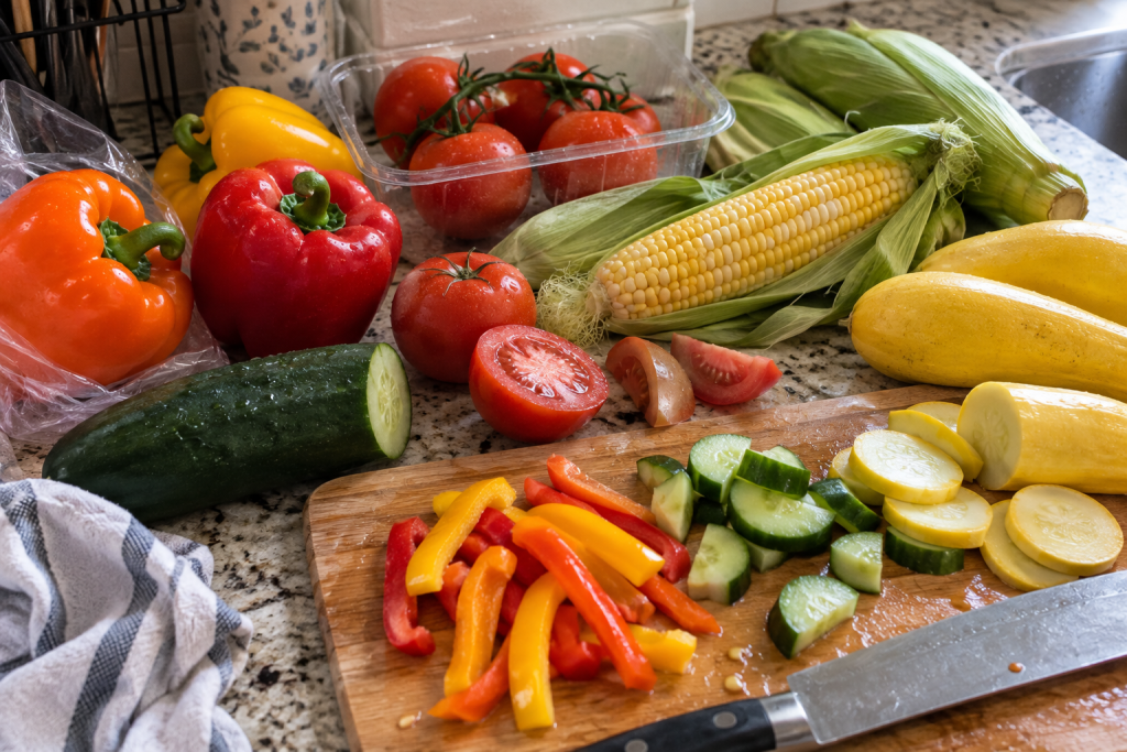 Fresh bell peppers, tomatoes, cucumber, summer squash, zucchini, and sweet corn being prepared on a kitchen counter