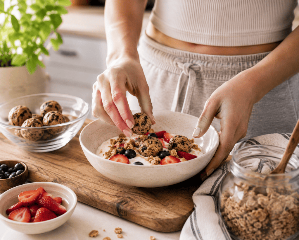 Hands placing Fit Five Meals Protein Bites into a Greek yogurt parfait topped with fresh strawberries, blueberries, and granola on a wooden countertop