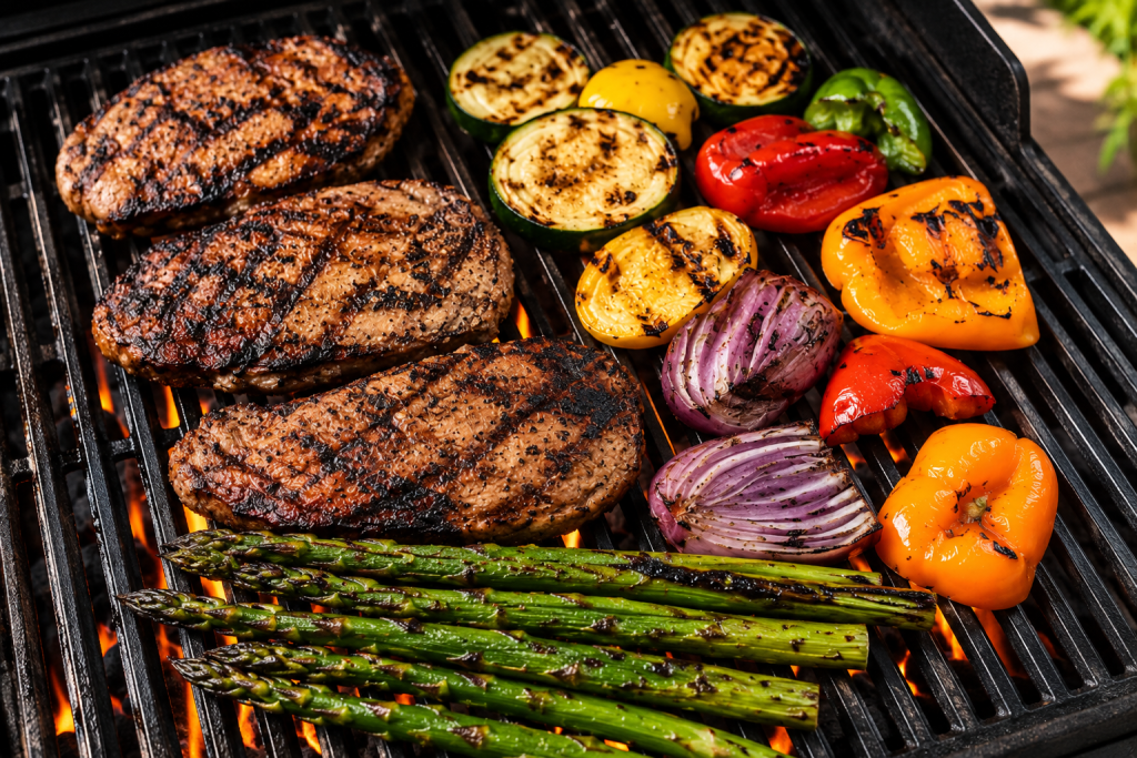 Grilled steaks with asparagus, zucchini, bell peppers, red onions, and other seasonal vegetables cooking over an outdoor grill.