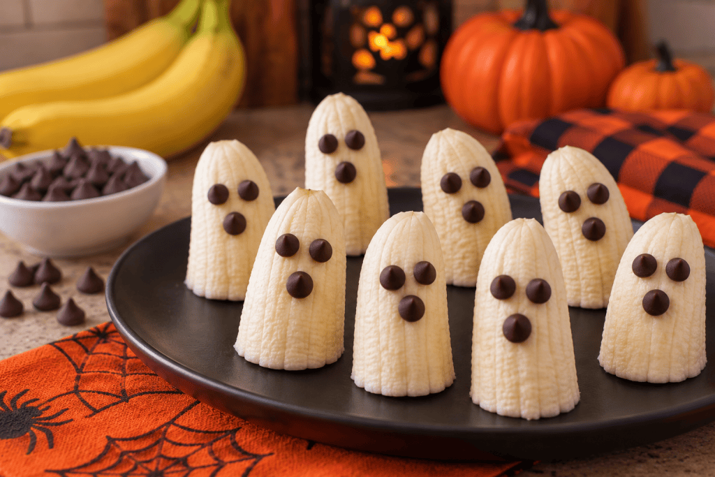 Boo-nana ghost treats made with peeled banana halves decorated with chocolate chip eyes and mouths, arranged on a black plate with festive Halloween decorations.